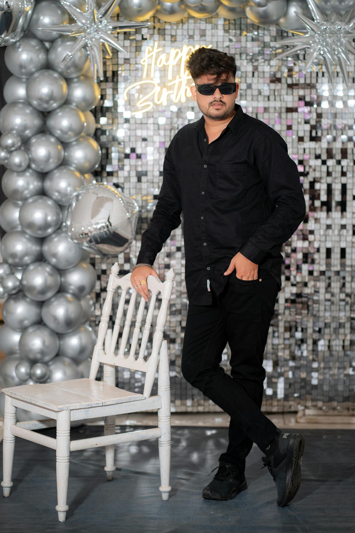 A stylish man poses with a chair at a birthday celebration featuring a silver theme with balloons and decor.